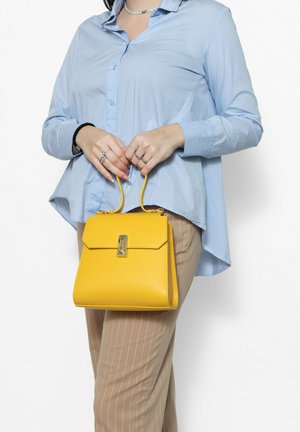 Borsa a mano in pelle gialla con un design strutturato, hardware in tonalità oro e manici corti, tenuta contro una camicia azzurro chiaro e pantaloni beige a righe.