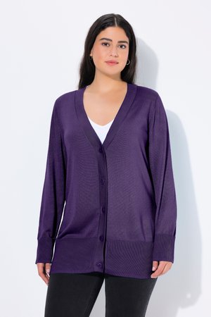 Kvinde iført en lilla cardigan med knapper over en hvid top og sorte bukser, stående mod en ensfarvet hvid baggrund.