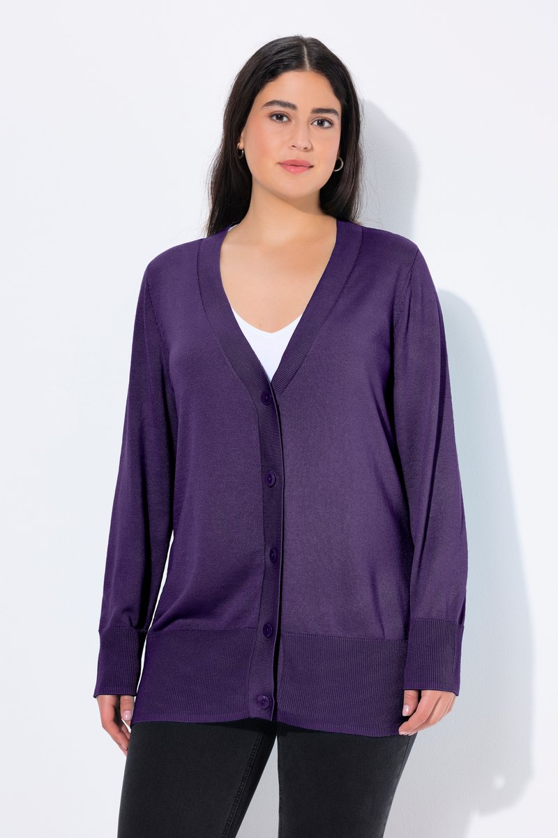 Donna che indossa un cardigan viola con bottoni sopra una maglietta bianca e pantaloni neri, in piedi davanti a uno sfondo bianco semplice.