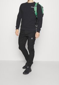 Musta pitkähihais sweatshirt, jossa on logo rinnassa, yhdistetty mustiin kapeneviin housuihin, joissa on brändätty päällekkäin, käytetään vihreän reppun kanssa.