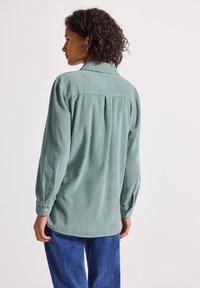 Donna con capelli ricci che indossa una maglietta a maniche lunghe a coste di colore verde chiaro e jeans blu, in piedi e rivolta di spalle rispetto alla fotocamera.