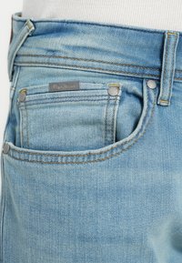 Ανοιχτό μπλε τζιν παντελόνι με ξεθωριασμένο φινίρισμα, που διαθέτει δύο μπροστινές τσέπες, ορατές ραφές και μια μικρή μεταλλική ετικέτα με λογότυπο στη μέση.