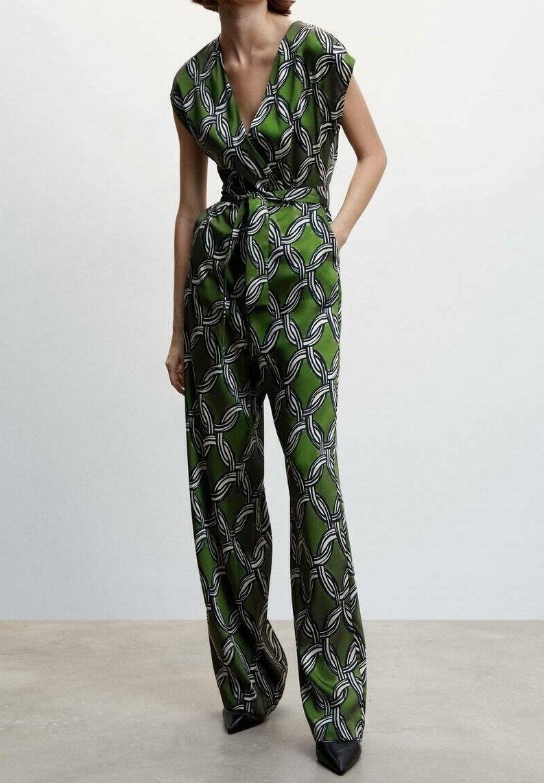 Femme portant une combinaison verte avec un motif chaîne noir et blanc, un décolleté en V, une taille cintrée et un pantalon à jambes larges, debout sur un sol uni.