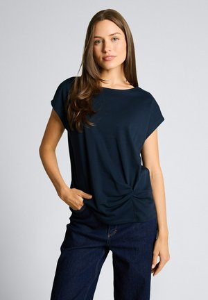 Vrouw met lang bruin haar, gekleed in een marineblauw kortemouwshirt met een strikdetail aan de zijkant en donkerblauwe spijkerbroek, staande met één hand in haar zak.