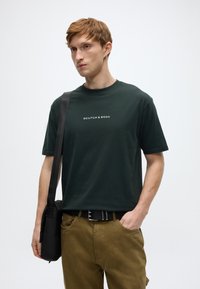 Σκούρο πράσινο βαμβακερό μπλουζάκι με στρογγυλή λαιμόκοψη και λευκό λογότυπο κειμένου. Συνδυασμένο με μπεζ παντελόνια και μαύρο τσαντάκι ταχυδρόμου.