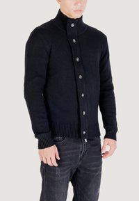 Uomo che indossa un cardigan lavorato a maglia nera con bottoni e un colletto alto, insieme a jeans neri sbiaditi, in piedi davanti a uno sfondo semplice.