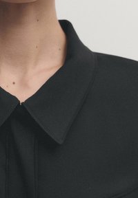 Zwarte kraagshirt met een gladde stoftextuur, voorzien van een puntige kraag en subtiele stiksels langs de randen.