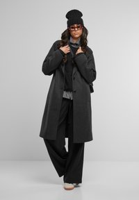 Cappotto oversized grigio scuro in lana, berretto nero a maglia, maglione nero, camicia a righe, pantaloni neri a gamba larga, occhiali cat-eye rossi, sneakers bianche.
