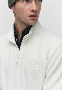 Witte fleece trui met een rits in de hals, geribbelde kraag en een gestructureerd oppervlak. Heeft een subtiele logo op de borst en een donker shirt eronder.