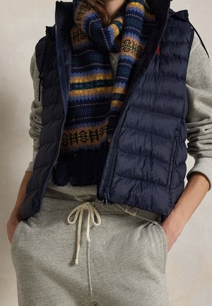 Persona con pantalones deportivos grises y sudadera con las manos en los bolsillos, que lleva un chaleco acolchado azul marino y una bufanda multicolor con estampado.