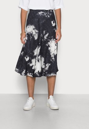 Kobieta ubrana w czarno-białą spódnicę midi tie-dye, białe trampki i biały krótki top, stojąca na jednolitym tle.