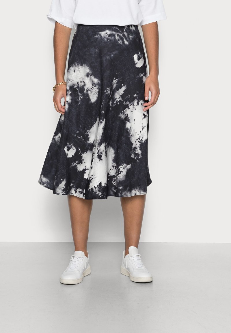 Femme portant une jupe midi tie-dye noir et blanc, des baskets blanches et un haut blanc à manches courtes, debout devant un fond uni.