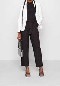 Svarta vida byxor med ett snörliv, kombinerade med en åtsittande svart topp och en vit blazer. Accessoarer inkluderar en liten silverhandväska och platåsandaler.