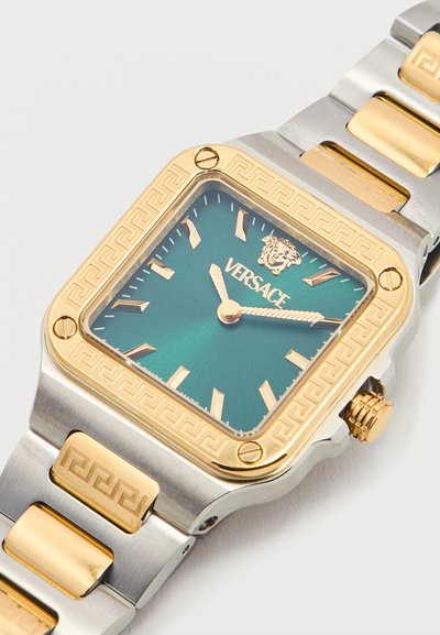 Τετράγωνο ρολόι χειρός Versace με δίσκο σε πράσινο μπλε χρώμα, χρυστούς δείκτες ώρας και ακροβατικά, χρυσό στεφάνι με μοτίβο ελληνικού κλειδιού και δίχρωμο βραχιόλι από ασημί και χρυσό μέταλλο.