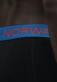Κοντινό πλάνο ζώνης μαύρου ρούχου με μπλε ταινία που φέρει έντονο πορτοκαλί κείμενο "NORWA" και ορατές ραφές στο ύφασμα.