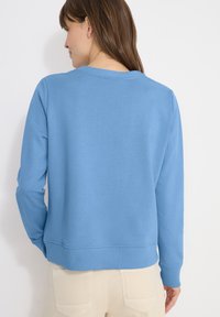 Femme aux cheveux bruns portant un pull bleu clair uni et un pantalon beige, vue de dos sur un fond blanc.
