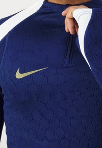Navy athletic top with a hexagonal texture, white accents on shoulders, gold Nike logo on chest, and a half zipper at the neck.