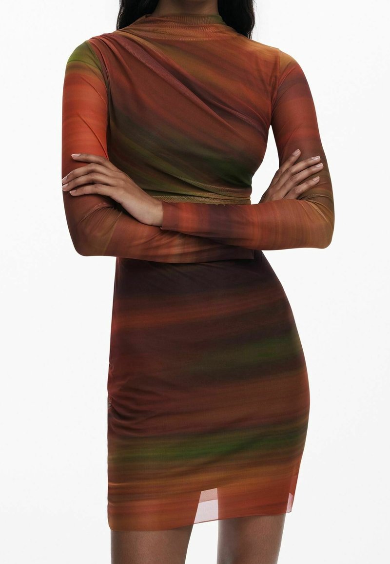 Femme portant une robe ajustée à manches longues multicolore avec des tons orange, vert et marron, bras croisés sur la poitrine.