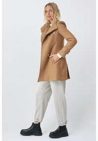 Abrigo marrón de doble botonadura con solapa, bolsillos frontales y cremalleras decorativas, combinado con pantalones gris claro y botas negras gruesas.