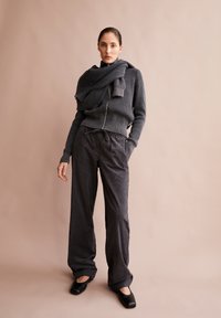 Donkergrijze geribbelde trui met rits aan de voorkant en oversized kraag, gecombineerd met brede corduroy broek en zwarte platte schoenen. Effen achtergrond.