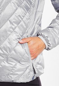 Srebrna ocieplana kurtka z pikowanym wzorem, boczną kieszenią zamykaną na suwak oraz elastycznymi mankietami, wykonana z gładkiego, lekkiego materiału.