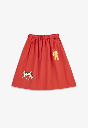 Niña con cabello largo trenzado, vistiendo una camiseta naranja, falda roja con estampados de vaca y granjero, calcetas rosas a rayas y zapatos rojos sin cordones.