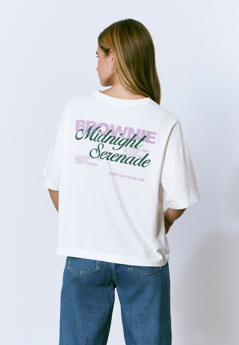 Mujer con cabello largo que lleva una camiseta blanca de gran tamaño con el texto "Serenata de Medianoche" y jeans azules, de espaldas frente a un fondo liso.