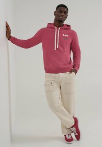 Rózsaszín kapucnis pulóver húzózsinórokkal, logóval és zsebrészletekkel. Krémszínű cargo nadrággal és piros tornacipővel párosítva.