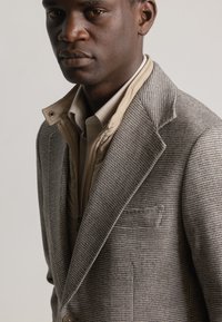 Blazer marrón de espiga con solapas en pico, dos botones y un bolsillo en el pecho, superpuesto a una camisa beige con cuello y cierre de cremallera en el frontal.