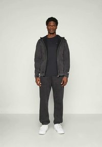 Giacca nera zip in tessuto testurizzato, abbinata a una t-shirt nera e pantaloni sportivi scuri; sneakers bianche; silhouette semplice e atletica.