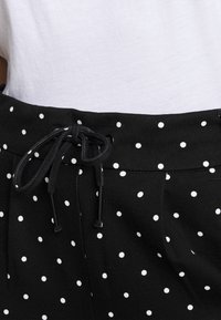 Calças de tecido preto com bolinhas brancas, apresentando uma cintura com cordão ajustável e dois bolsos laterais. Textura suave, design casual.