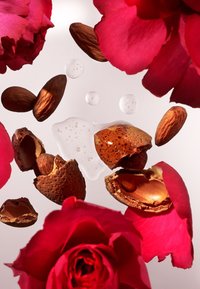 Mandorle, spezzate e intere, circondate da vivaci petali di rosa rossa. Bollicine e gocce di liquido appaiono su uno sfondo chiaro.