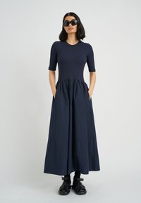 Marineblaues Maxikleid mit passgenauem, geripptem Oberteil und gerafftem Rock. Mit kurzen Ärmeln und Seitentaschen. Das Model trägt schwarze Sonnenbrillen und Schuhe.
