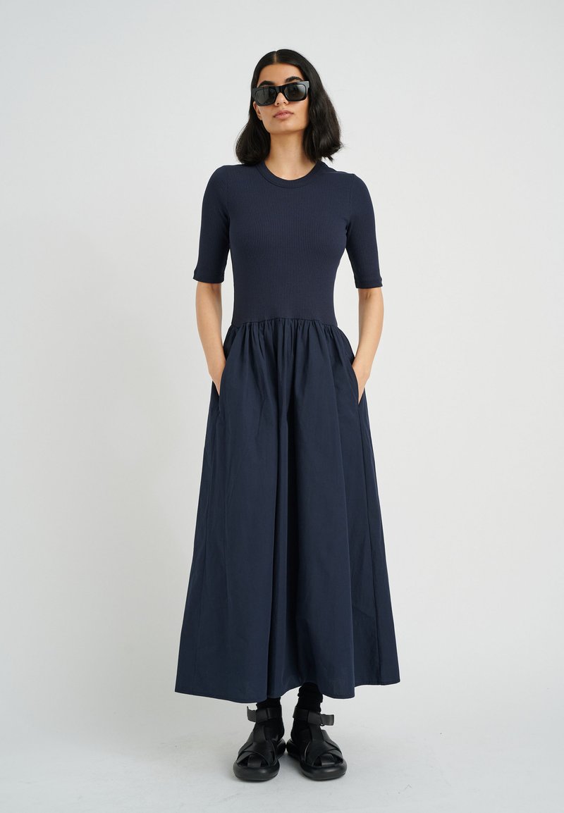 Marineblaues Maxikleid mit passgenauem, geripptem Oberteil und gerafftem Rock. Mit kurzen Ärmeln und Seitentaschen. Das Model trägt schwarze Sonnenbrillen und Schuhe.