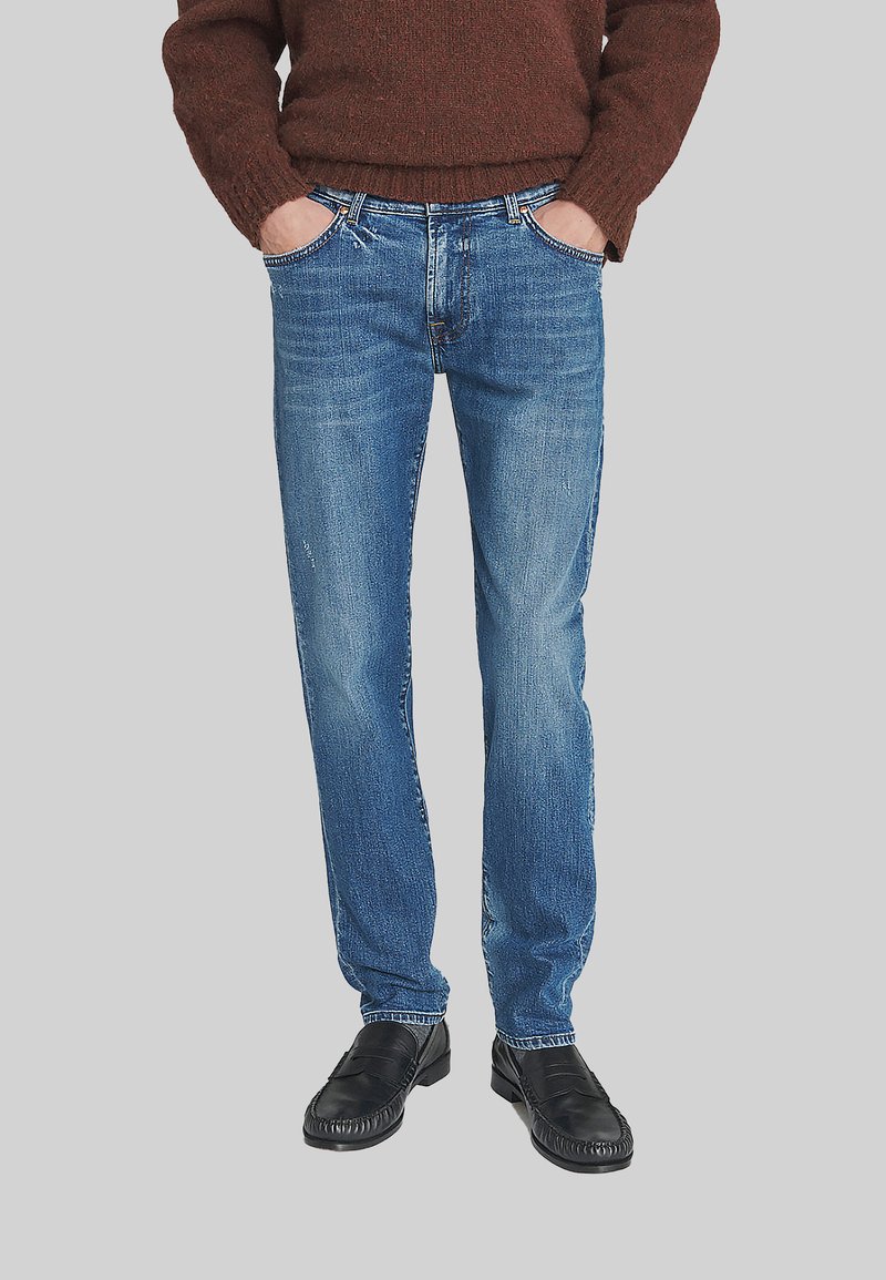 Uomo che indossa jeans blu, mocassini neri e un maglione marrone, con le mani nelle tasche davanti, in piedi su uno sfondo grigio uniforme.