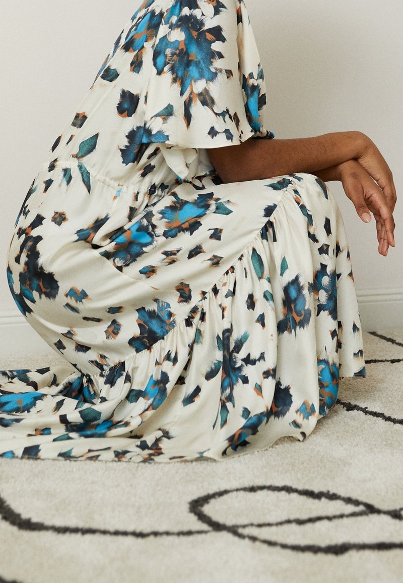 Personne agenouillée sur un tapis à motifs portant une robe blanche avec un imprimé floral abstrait bleu, noir et marron.
