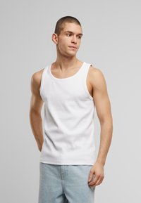 White sleeveless tank top in cotton, featuring a rounded neckline and a fitted design. Paired with light blue denim jeans.