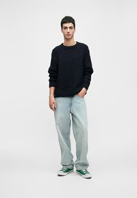 Mornarska pletena majica z okroglo izreza, v kombinaciji s svetlo modrimi ravnimi hlačami in zelenimi ter belimi športnimi copati.