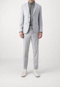 Suit grigio chiaro su misura con un blazer strutturato a due bottoni, abbinato a una camicia verde scuro con bottoni e scarpe da ginnastica bianche.