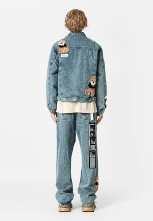 Chaqueta de mezclilla y pantalones en azul claro. La chaqueta presenta diseños bordados de osos y etiquetas en la manga. Los pantalones tienen gráficos en el costado y acentos de oso.