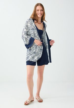 Femme souriante portant une combinaison bleu, une veste style kimono à motifs, un collier de perles et des sandales blanches sur un fond clair uni.
