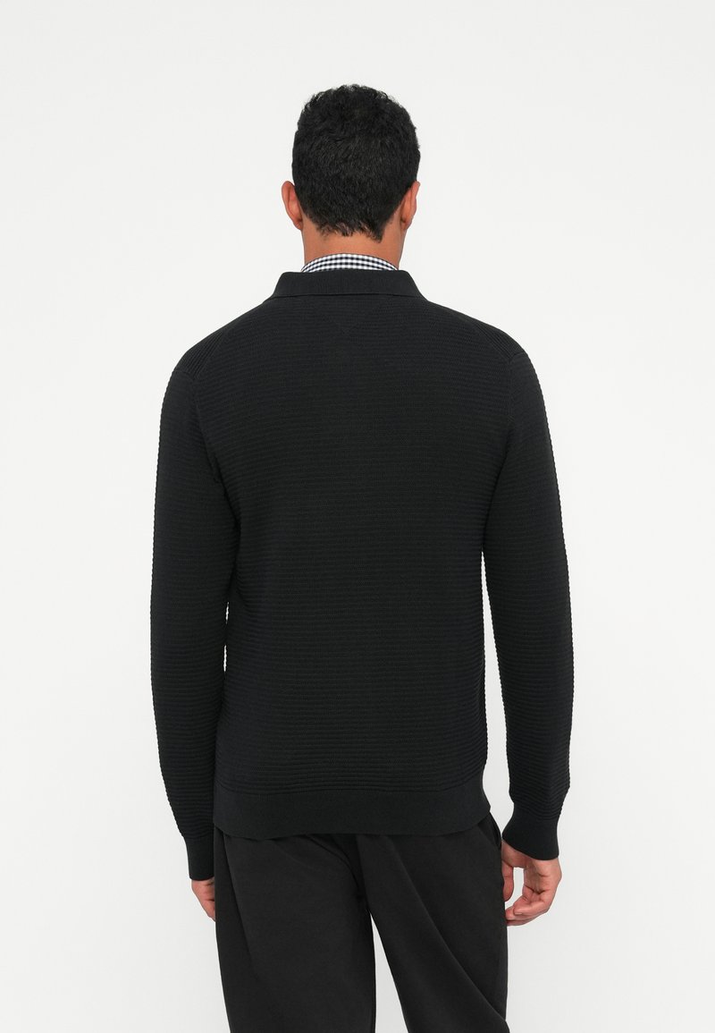Black textured sweater with a ribbed pattern, collared neckline, and long sleeves, styled over dark trousers.