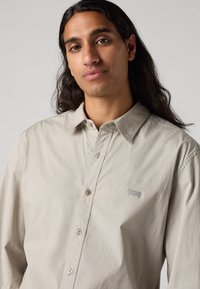 Chemise boutonnée à manches longues de couleur beige clair, avec un col pointu, arborant un petit logo brodé sur la poitrine et des poignets boutonnés.