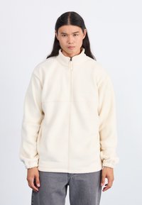 Karamelli fleece takki, jossa on korkea kaulus, puolivälin vetoketju ja elastiset hihat, sekä sileä pinta ja minimalistinen muotoilu.