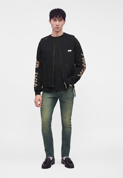 Jeune homme portant une veste noire à manches graphiques, un jean slim vert délavé et des mocassins noirs à pampilles, debout avec une main dans la poche.