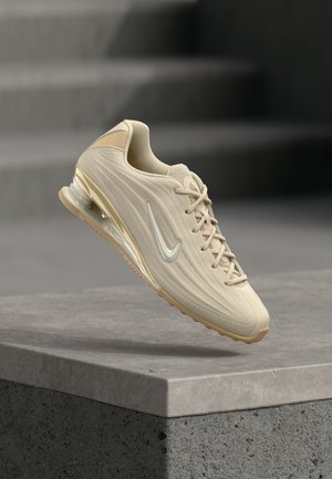 Beige Nike sneaker with textured upper, white swoosh logo, laces, and air-cushioned sole floating above a concrete ledge.