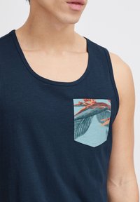 Námornícky modré tielko s okrúhlym výstrihom a prednou vreckom zdobeným farebným vzorom listov na svetlomodrom látku.