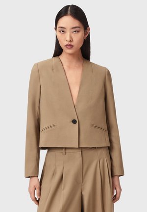 Vrouw draagt een beige cropped blazer met een enkele zwarte knoop, gecombineerd met bijpassende, hoog getailleerde, geplooide broek, tegen een effen achtergrond.