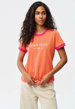 Kobieta z długimi kręconymi włosami ubrana w pomarańczowy, pasiasty T-shirt z napisem "Reach Beach" i kremowe spodnie, stoi na tle jasnego, jednolitego tła.