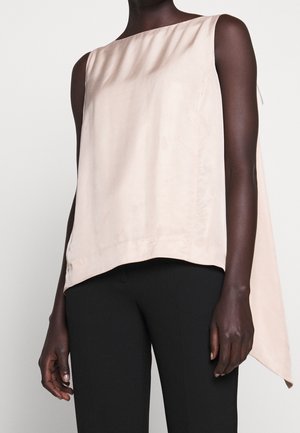 Osoba o ciemnej karnacji ubrana w bezrękawnik w jasnobeżowym kolorze o asymetrycznym kroju z dłuższym tyłem, zestawiona z dopasowanymi czarnymi spodniami, stojąca na neutralnym tle.